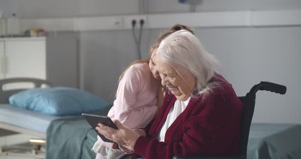 Little Girl Hugging Disabled Grandmother in Wheelchair Using Digital Tablet in Nursing Home