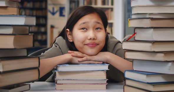 Happy Asian School Student Sitting in Library Leaning on Pile of Books