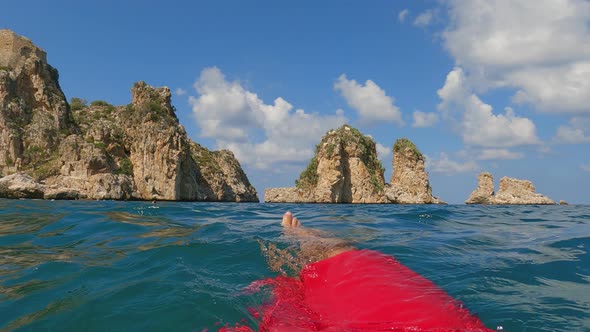Fpv of male legs and feet relaxing while floating on sea water with Scopello Stacks or Faraglioni in