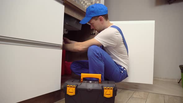 plumber in overalls and white T-shirt examines pipes in kitchen in apartment.