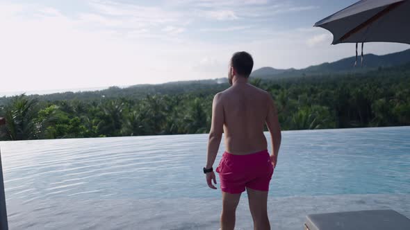 A Shirtless Caucasian Man Walks Up to the Swimming Pool in Swim Trunks to See a Picturesque Jungle