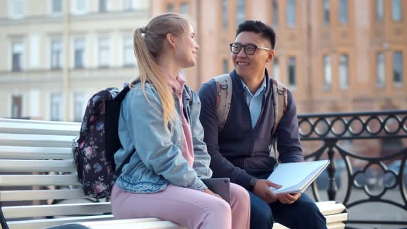 Happy Young Multiethnic Couple Sitting on Bench in University Campus.