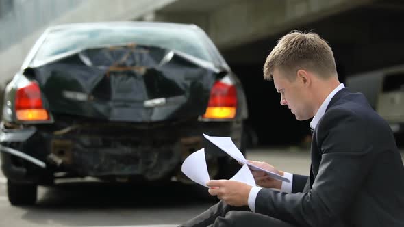 Male Driver Sitting on Road, Looking Through Insurance Contract After Car Crash