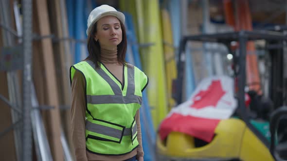 Serious Confident Beautiful Woman in Hard Hat Looking Around at Racks with Goods in Warehouse and