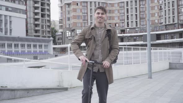 Portrait of Cheerful Adult Caucasian Man Standing on City Street with Scooter and Smiling at Camera