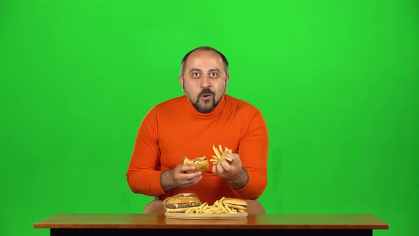 Caucasian Man at a Table with a Plate of Fast Food Enjoys Eating a Burger and French Fries, Green