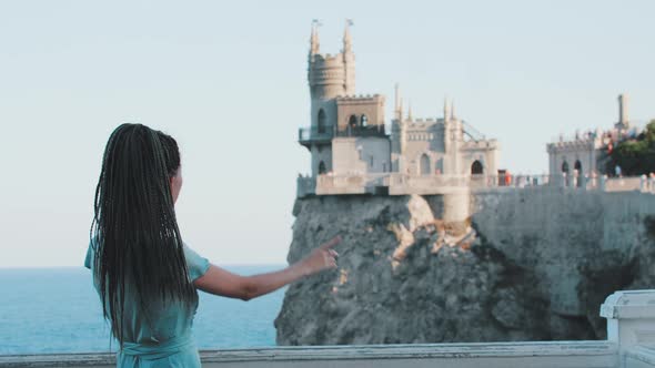 A Young Woman with Dreadlocks Stands on a Background of Sea and a Castle on the Edge Cliff Talking