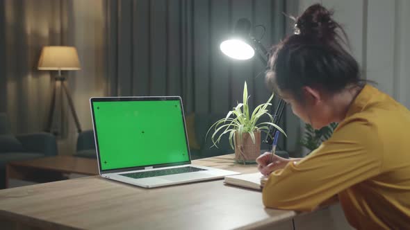Girl Works On A Laptop Computer With Mock-Up Green Screen, In The Background Evening At Home