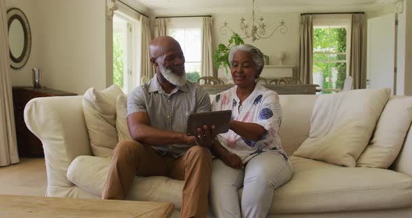African american senior couple looking at each other and smiling while using digital tablet at home