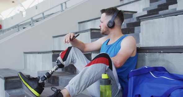 Caucasian disabled male athlete with prosthetic leg sitting, wearing headphones, using smartphone