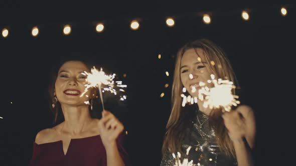 Two Multiracial Girlfriends in Plush Dresses Dancing in Dark at Party Holding and Waving Sparklers