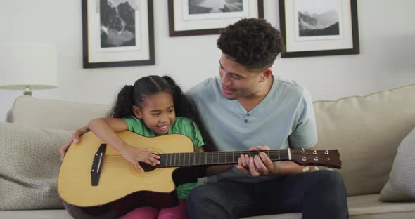 Happy biracial father and daughter sitting on sofa playing guitar