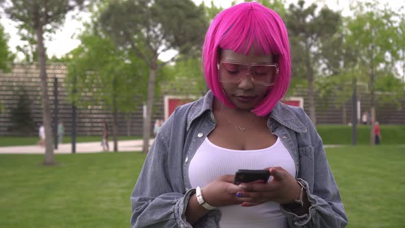 A Young Stylish Black Woman with Pink Hair Uses Her Smartphone Outdoors