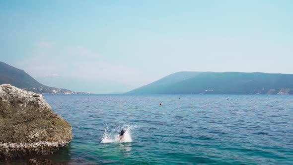 Adult woman jumping off from a rock into the sea water on a clear day. Static, slow motion shot