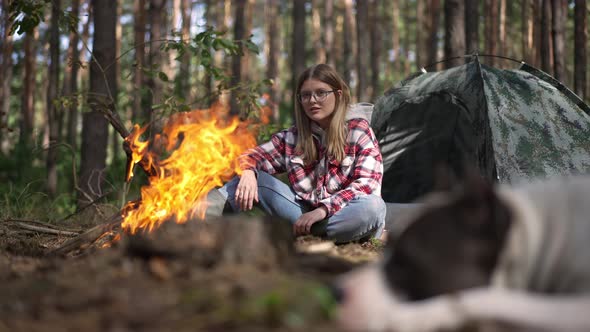 Caucasian Young Woman Admiring Bonfire in Forest Sitting at Tent Talking to Dog