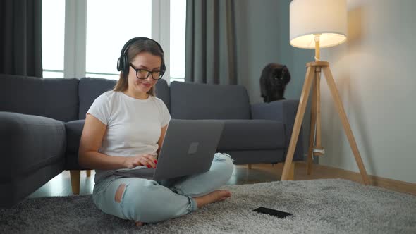 Casually Dressed Woman with Headphones is Sitting on Carpet with Laptop and Works Remotely in Cozy