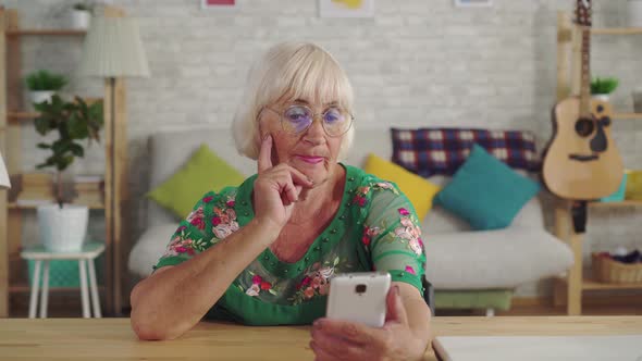 Thoughtful Elderly Woman Uses a Smartphone Sitting at the Table in the Living Room