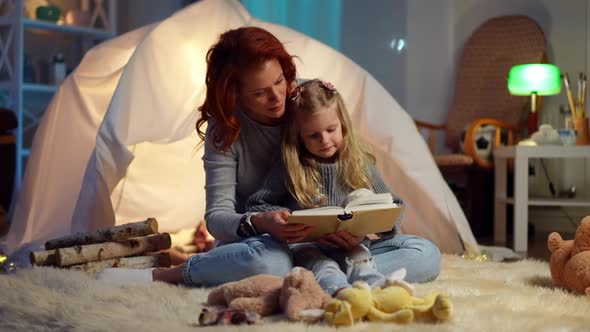 Wide Shot Portrait of Beautiful Caucasian Mother Reading Book with Curios Pretty Daughter Sitting at