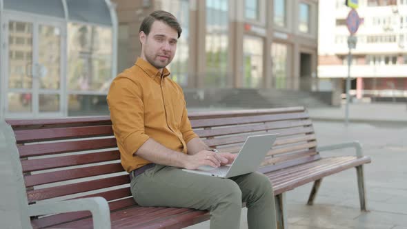 Young Man Shaking Head in Approval While using Laptop Sitting Outdoor on Bench