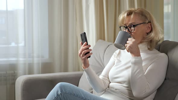 Mature Woman Sips Coffee and Reads News Online Via Phone