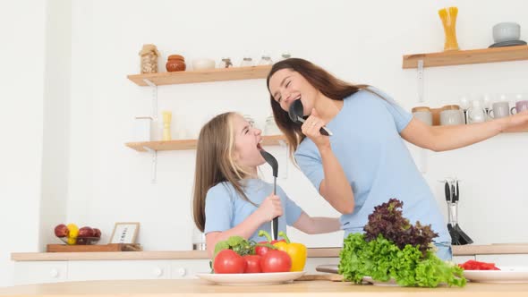 Young Mother with Happy Young Little Daughter Singing in Karaoke Holding Kitchen Spoon in Hands