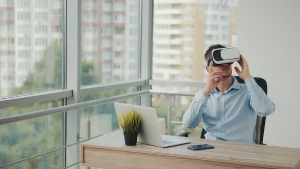 A Young Man Sitting at a Desk in the Office Uses Augmented Reality Glasses To Work on Business