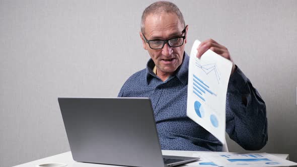 Elderly Man Checks Drawings Near Laptop at White Office Desk