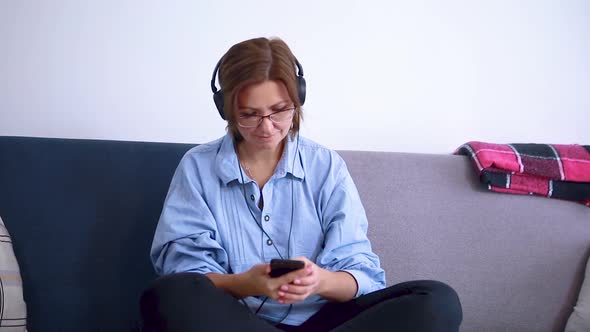Woman in glasses with headphones listens to music on a mobile phone while sitting on the couch
