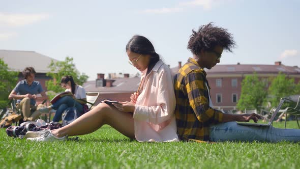 Side View of African and Asian Students Sitting Back to Back and Study at Campus Lawn