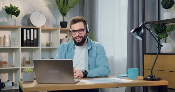 Bearded Guy in Headphones Holding Video Chat with Friend or Colleague Sitting in Home office