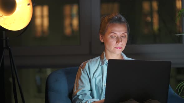 Woman is Sitting in the Armchair and Working on a Laptop at Night