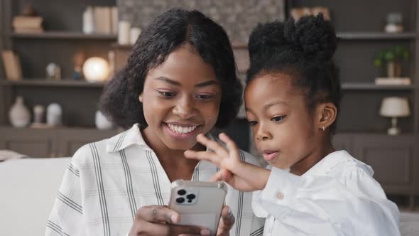 Cute Little African Kid Child Daughter Having Fun with Multiracial Mum at Home Using Smart Phone