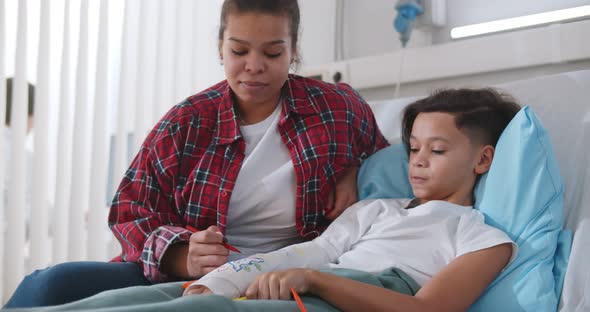Black Mother Visiting Son Lying with Broken Arm in Bed in Hospital Ward