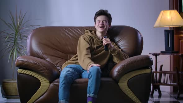Cheerful Caucasian Boy Laughing Out Loud As Sitting on Armchair with Remote Control. Joyful Teenager