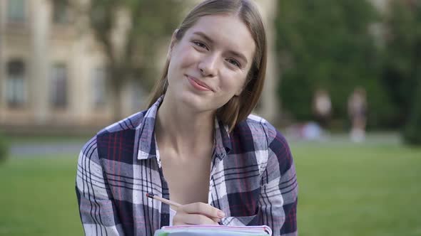 Attractive Female Student Sitting on The Grass and Writing in The Textbook