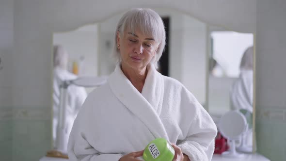 Sad Caucasian Middleaged Greyhaired Woman Standing in Bathroom with Aloe Cream Looking at Camera
