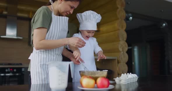 Mom in Apron with Her Son Dressed in Chef's Uniform Kneading Dough for Apple Pie in the Kitchen