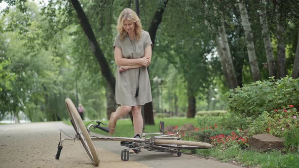 Wide Shot of Sad Middle Aged Woman Rubbing Injured Elbow and Kicking Bicycle Lying on Park Alley