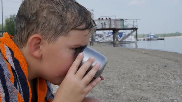 Caucasian Teenage Boy with a Wet Body and Head Sits on the Bank of the River Wrapped in a Towel