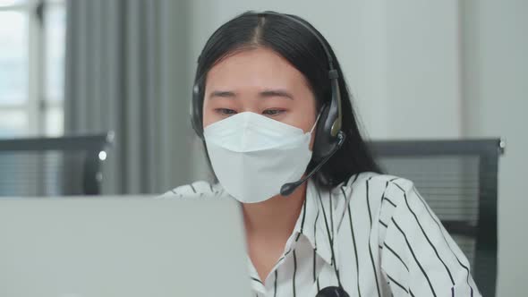 Close Up Of An Asian Woman Call Centre Agent Wearing Headset, Mask While Speaking To A Customer