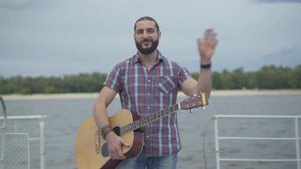 Positive Caucasian Man Standing with Guitar on Embankment and Waving. Portrait of Happy Young