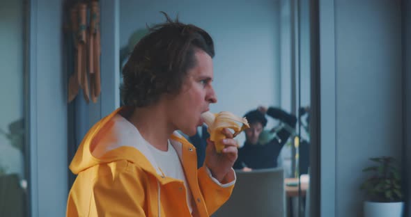 Young man takes a bit from a banana and stands up