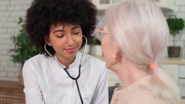 Afro American Woman Doctor Listens to Breath of Senior Woman Using Stethoscope While Sitting on Sofa