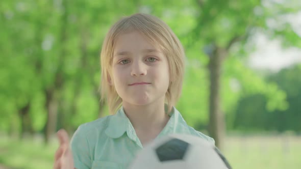 Close-up of Charming Blond Caucasian Boy with Green Eyes Posing with Soccer Ball in Sunny Park