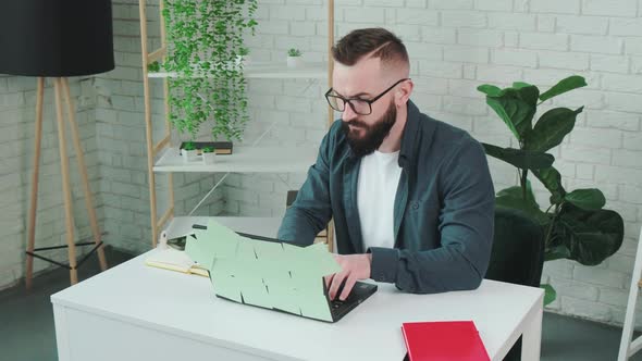 Bearded Man Types at the Laptop and Write Notice on the Paper Notebook