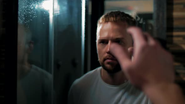 Man wipes a misted mirror with his hand and look at his reflection in the bathroom