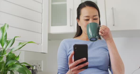 Happy asian woman drinking coffee and using smartphone in kitchen
