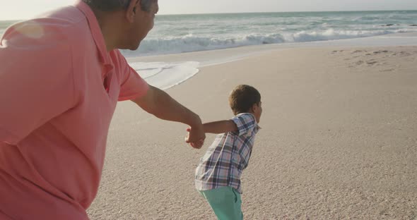 Happy hispanic grandfather and grandson running on beach at sunset