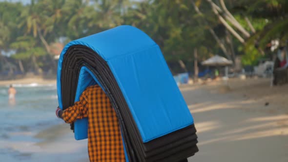 Dark Skinned Man Holds Mats on Head and Walks Along Beach