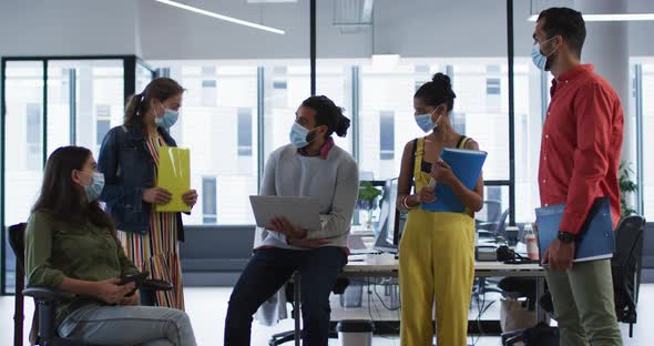 Diverse group of work colleagues wearing face mask holding laptop, tablet and documents talking